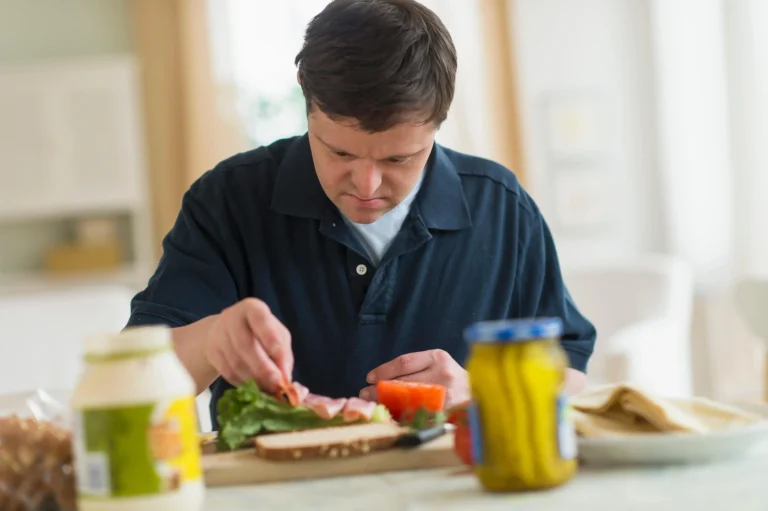 A person making a sandwich at a table, placing ingredients like lettuce, tomato, and ham on bread, with jars of mayonnaise and pickles nearby.